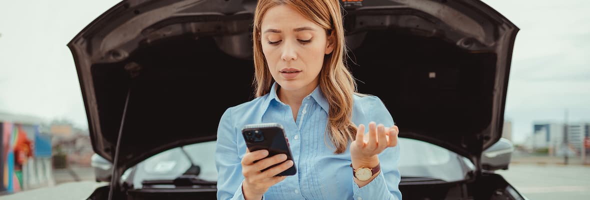 A frustrated woman calls roadside assistance for help in front of her broken down car's open bonnet.