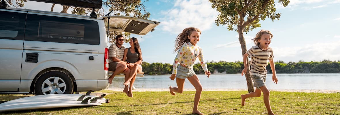 Two children run along a lakeside while their parents watch from the back of a campervan