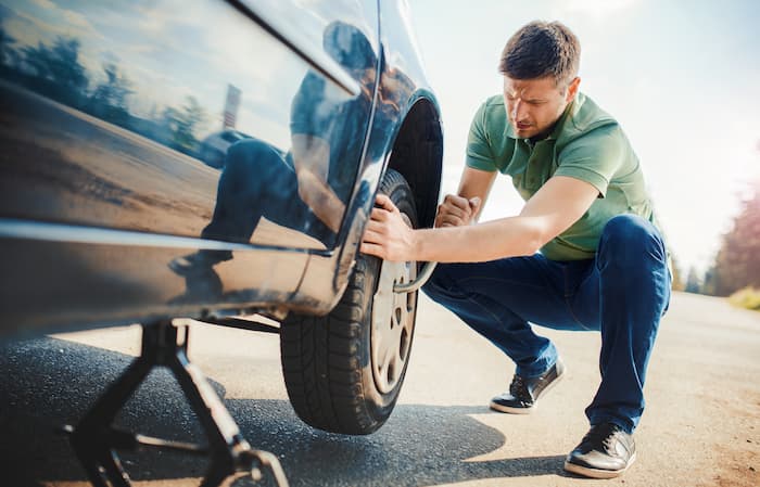 A man kneels down and uses a wrench to change a flat tyre on the side of the road.