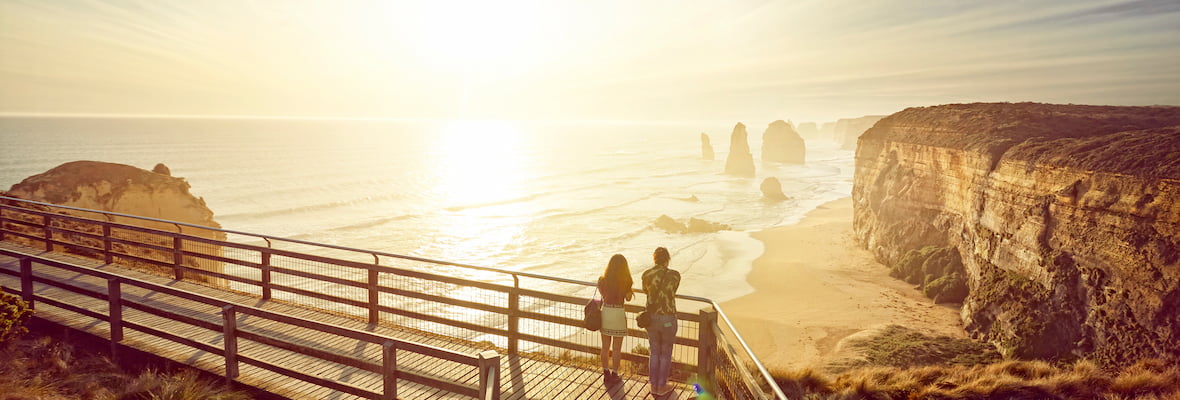 Couple stand on boardwalk looking at great ocean road at sunrise