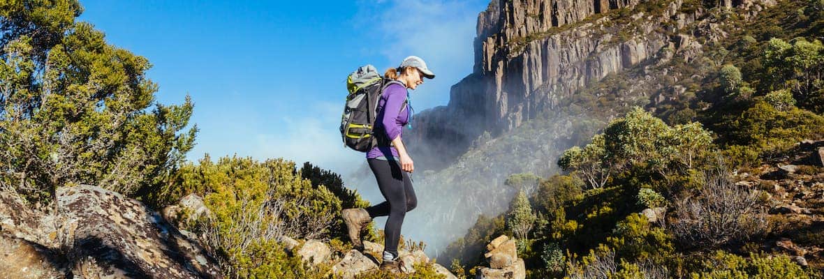 A woman hikes the rocky trail through green shrubbery to the top of Mount King William I in Tasmania, Australia.