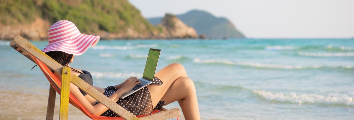 woman sitting on deck chair on tropical beach while working on laptop